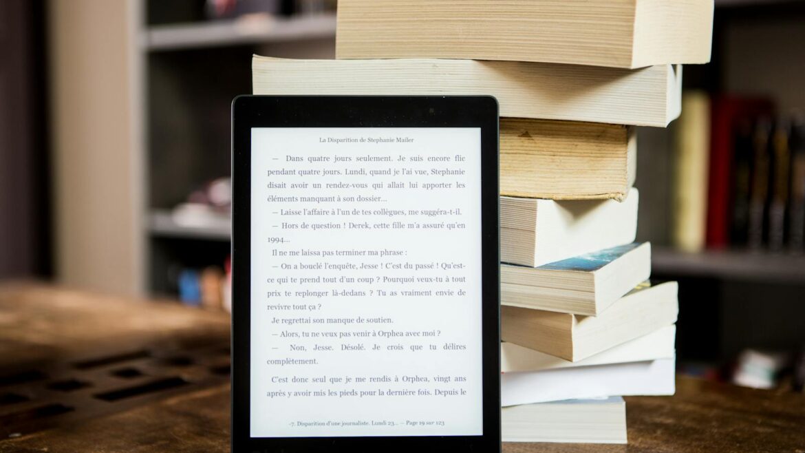 E-reader with text displayed next to a stack of physical books on a wooden table.